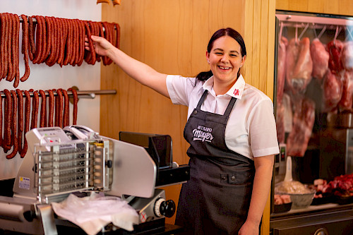 Das Bild zeigt eine Mitarbeiterin, die vor einer Schneidemaschine steht und ihre Hand nach einer darüberliegenden Wurst ausstreckt. Dabei lächelt sie in die Kamera.