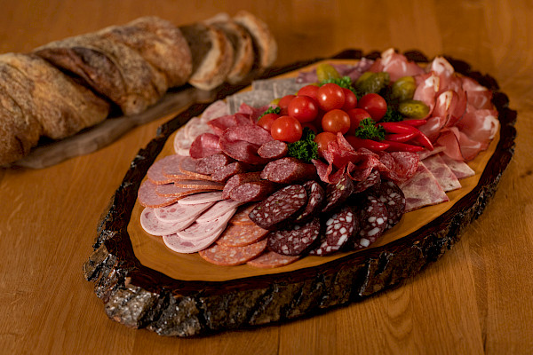 Das Bild zeigt eine Wurstplatte auf einem Holzbrett präsentiert. Die Platte steht auf einem Tisch. Im Hintergrund liegt ein zum Teil aufgeschnittenes Brot auf einem Holzbrett.