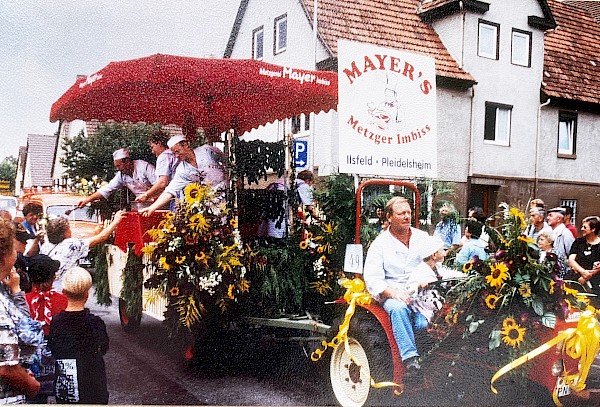 Holzmarkt im Jahre 2000. Ein Traktor zieht einen Anhänger, der mit Blumen geschmückt ist und auf welchem Mitarbeitende kleine Würste an die Zuschauer verteilen auf einem Umzug.