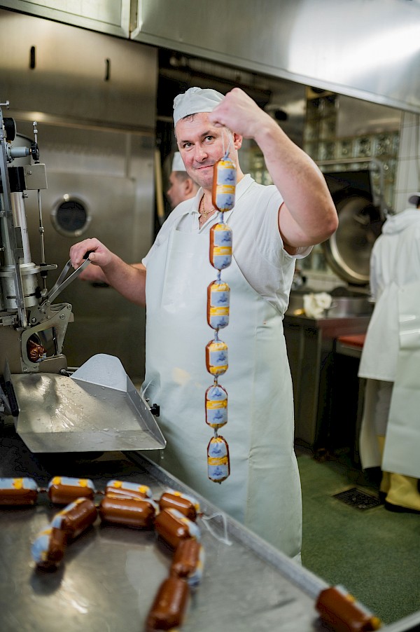Metzger hält eine Schlange Wurst in der Hand, steht an einer Maschine und schaut in die Kamera.