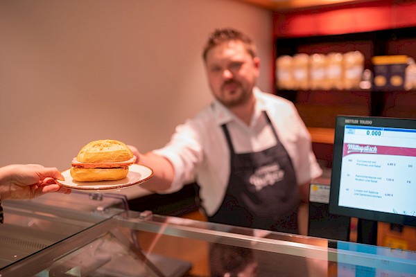 Verkäufer überreicht ein Teller mit einem Fleischkäsebrötchen.