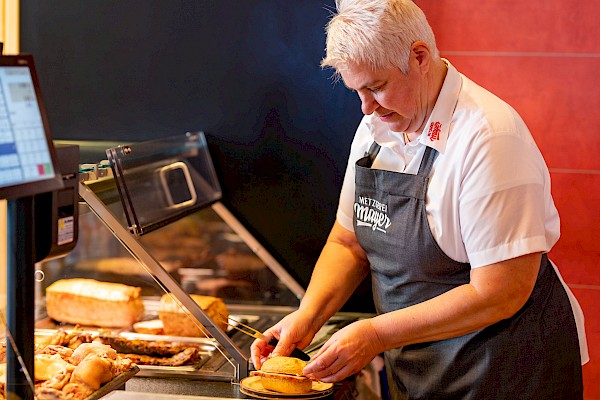 Verkäuferin steht an der Warmtheke und bereitet ein Fleischkäsebrötchen vor.