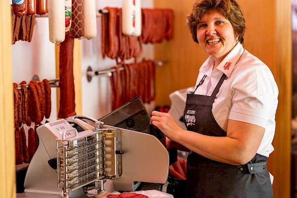 Eine Verkäuferin steht an der Wurstschneidemaschine und lächelt dabei in die Kamera.
