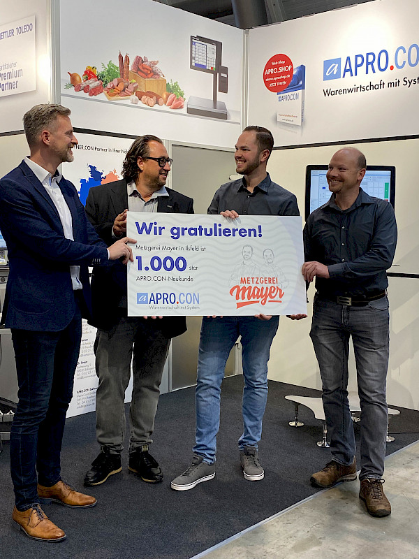 Tobias Fichtel und Frank Mayer halten gemeinsam mit zwei Mitarbeitern der Apro.Con Firma ein Plakat in die Höhe, auf welchem die Metzgerei Mayer als 1000 Kunde von AproCon ausgezeichnet wird.