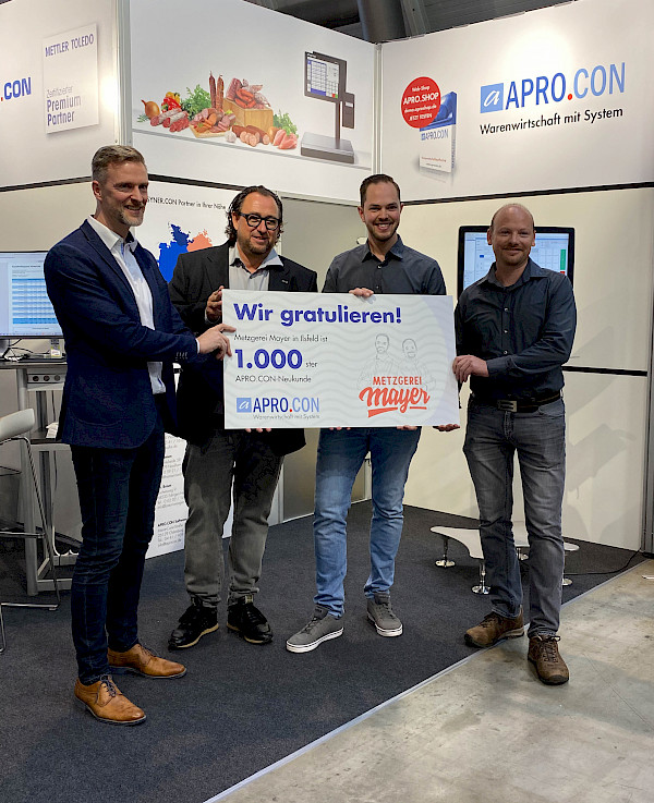 Tobias Fichtel und Frank Mayer halten gemeinsam mit zwei Mitarbeitern der Apro.Con Firma ein Plakat in die Höhe, auf welchem die Metzgerei Mayer als 1000 Kunde von AproCon ausgezeichnet wird. Alle schauen lächelnd in die Kamera.