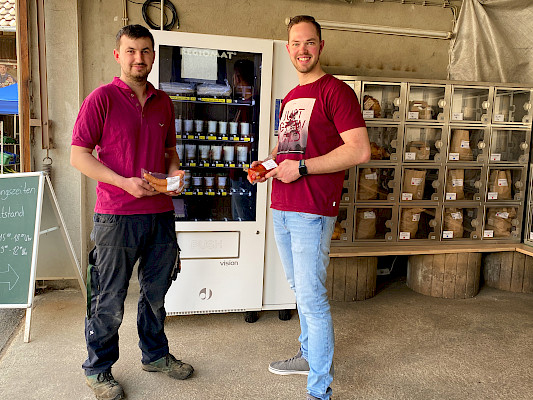 Tobias Fichtel und Andreas Obenland stehen vor einem Automat und halten Wurst- und Fleischwaren in der Hand, die man in diesem Automat kaufen kann.