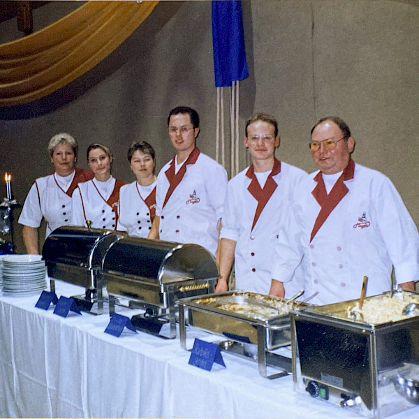 Das Bild zeigt Familie Mayer in jungen Jahren hinter einer Tischreihe, auf welcher ein Buffet aufgereiht ist.